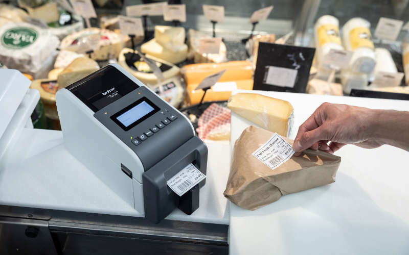 Label printer on the counter surrounded by cheese with a hand applying a label to a piece of cheese