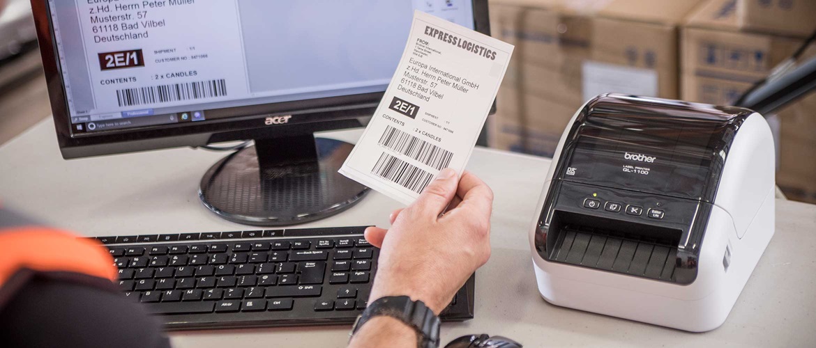 Brother QL-1100 desktop label printer on desk with person sat at desk holding label